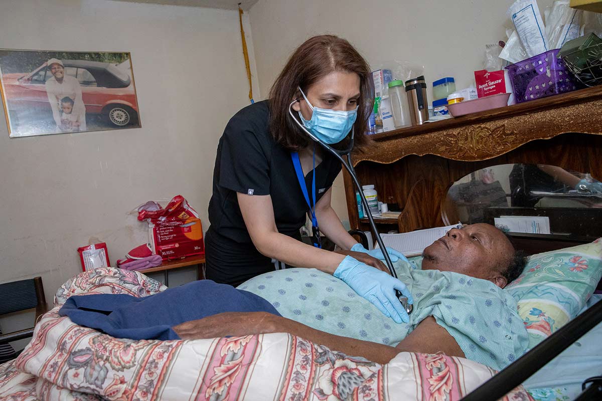 Woman in scrubs and mask using stethescope on chest of woman laying in bed