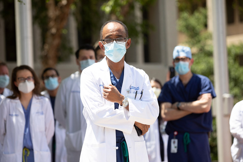 Black man with glasses, surgical mask, wearing white lab coat over scrubs, speaking