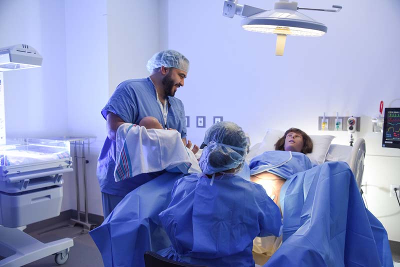 Man in scrubs holding dummy baby next to woman in scrubs while looking at dummy patient in hospital bed