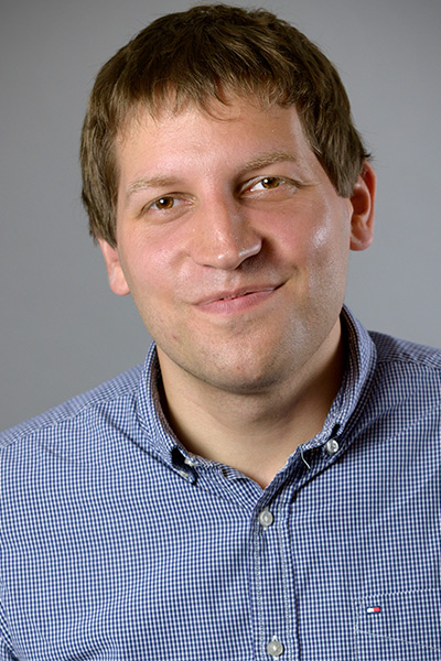 Man with dark hair, thin framed glasses, grey collared shirt wearing a white lab coat