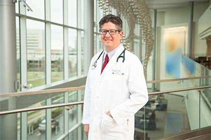 Dr. Blake Barker white man with dark hair, glasses, white lab coat and stethoscope on his neck standing in sunny glass atrium