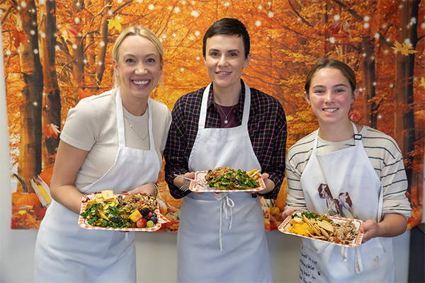 three ladies in aprons display holiday food in front of autumn leaves background