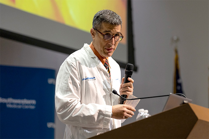 Man with salt-n-pepper hair and glasses wearing a lab coat speaking from a podium, holding a microphone.