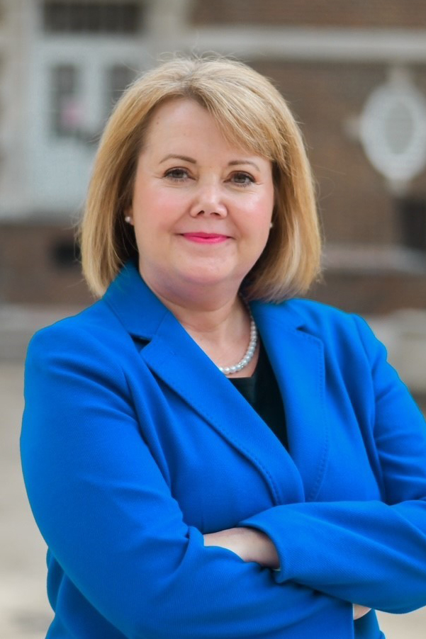 Smiling blond woman, wearing a blue suit and pearls, standing in front of a brick building.