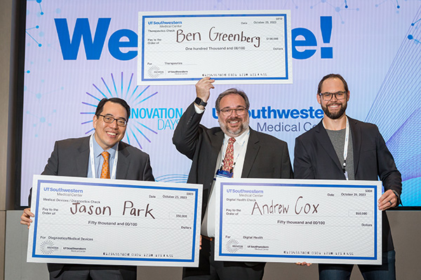 three men in gray suits smile and hold up large signs that look like checks showing their award winning amounts for research