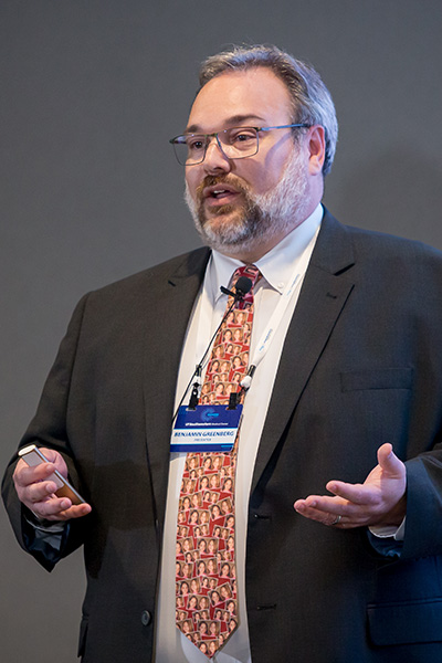 heavyset man with glasses and beard wears dark suit with red patterned tie; gestures as he speaks