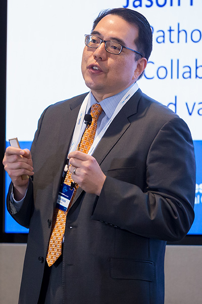 Asian man with dark hair and glasses gestures during presentation of slide in background