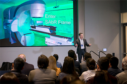 Man on a stage in front of a large screen, speaking to an audience.