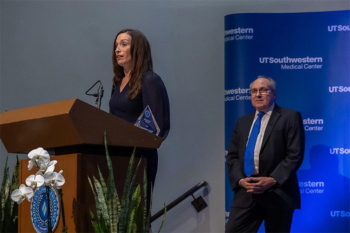 Dr. Herrera, woman with long dark hair, dressed in black, standing at a podium. Dr. Podolsky standing in background in front of a UTSW backdrop.