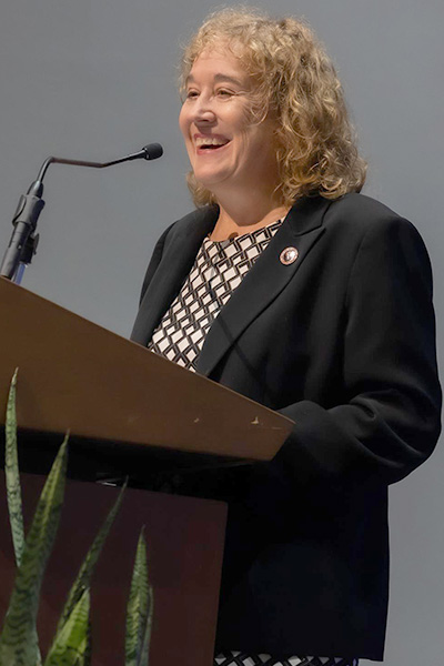 Dr. Kowalske, smiling woman with curly blond hair dressed in black, standing at a podium.