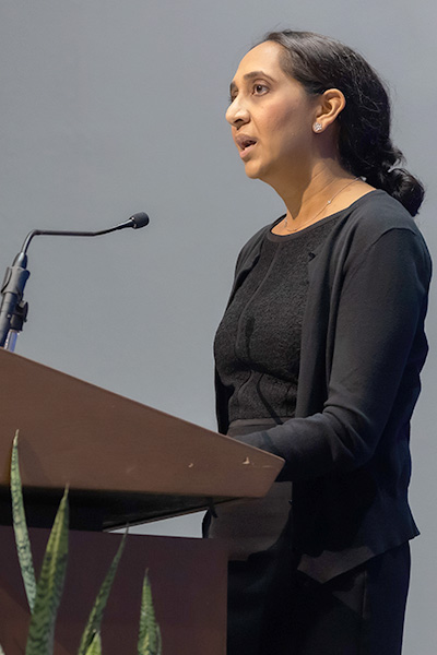 Dr. Patel, woman with long dark hair pulled back from her face, wearing black, standing at a podium.