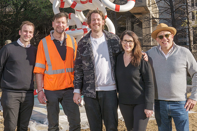 Four men and one woman pose outdoors in front of a statue made of round lifepreservers.