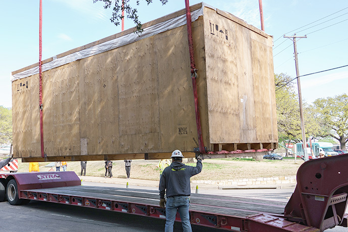 Crane placing very large crate on back of flat bed truck, with to workmen helping to guide it.