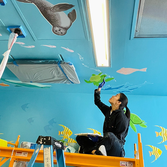 Dr. Chen sits on a scaffold to paint a turtle on the ceiling mural