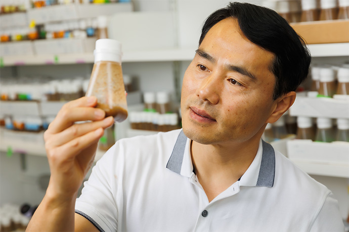 A man with dark hair wearing a white shirt examines the contents of the beaker he holds in his hand.