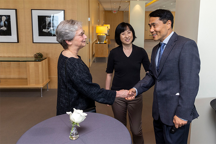 A woman with short gray hair shakes hands with a man with dark hair wearing a gray suit, as a woman with dark hair watches.