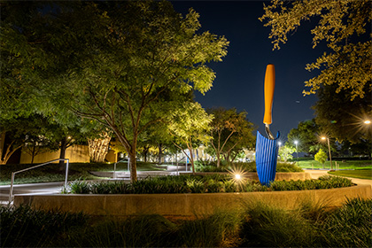 A night time view of a large blue and gold sculpture of a hand spade for gardening, partially embedded in the ground among trees.