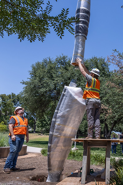 Two men in orange vests and hard hats work to lower a wrapped handle down onto the large trowel of the sculpture