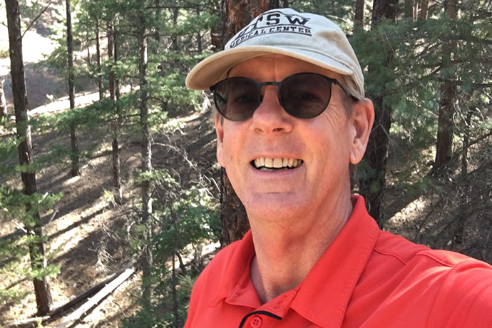 smiling man wearing UTSW baseball cap and sunglasses with pine forest behind him