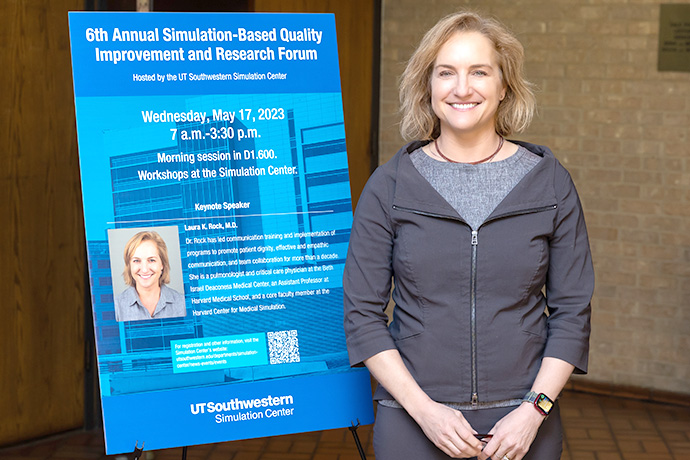Blond woman in gray jacket standing next to blue poster.