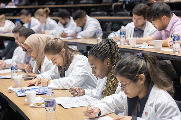 Men and women in lab coats sitting at rows of tables, writing in notebooks.