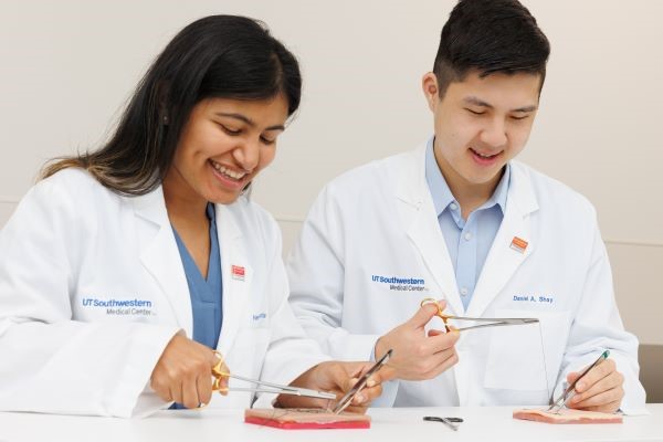 female and male med students in white coats sit together practicing sutures in simulation