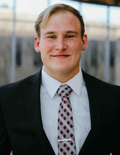 Smiling blond man wearing a dark suit and plaid tie.