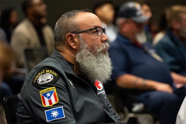 balding man with glasses and gray beard wears shirt displaying Combat Medic and Puerto Rico flag emblems
