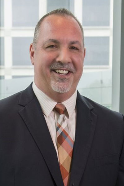 Smiling man with light hair and gray beard and mustache, wearing a dark gray suit, white shirt and plaid tie