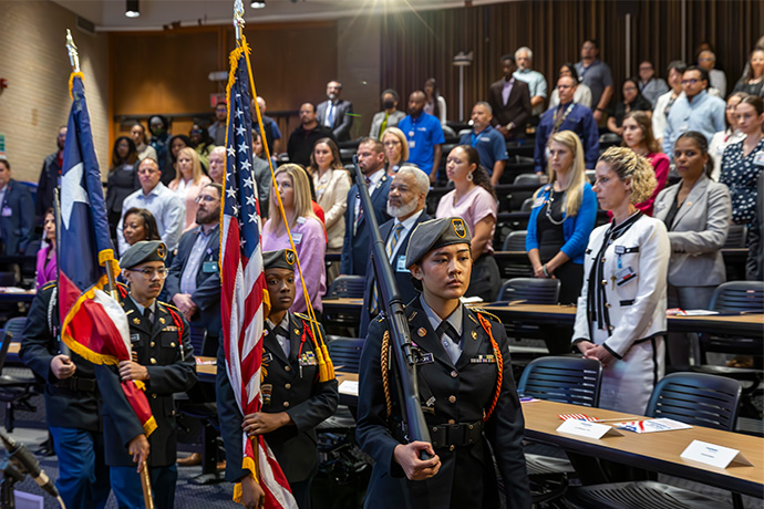 dozens of professionals stand in auditorium behind line of 3 young JROTC color guard members wearing green berets and uniforms, holding (from right) rifle, American flag and Texas state flag