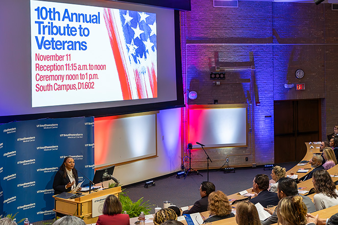 Dr. Nesbitt speaks from podium at front of auditorium; projector above her shows '10th Annual Tribute to Veterans November 11' title slide