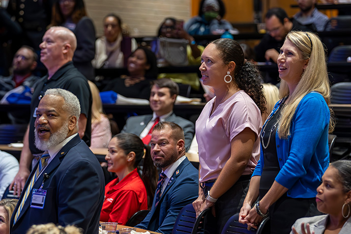 seated audience smiles during program as 4 veterans stand for recognition; from left: balding white man in polo shirt, black man with grey hair and beard, woman with long dark ponytail and hoop earrings, and woman with long blonde hair and blue cardigan