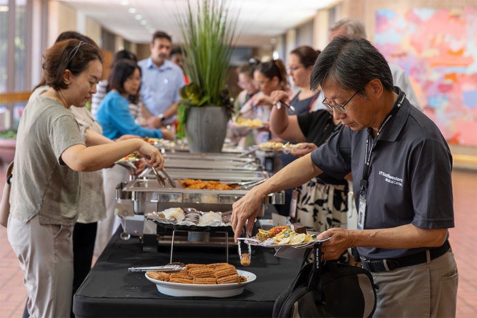 woman at left, man wearing UTSW polo shirt at right spoon food from buffet pans; more attendees in background also in line