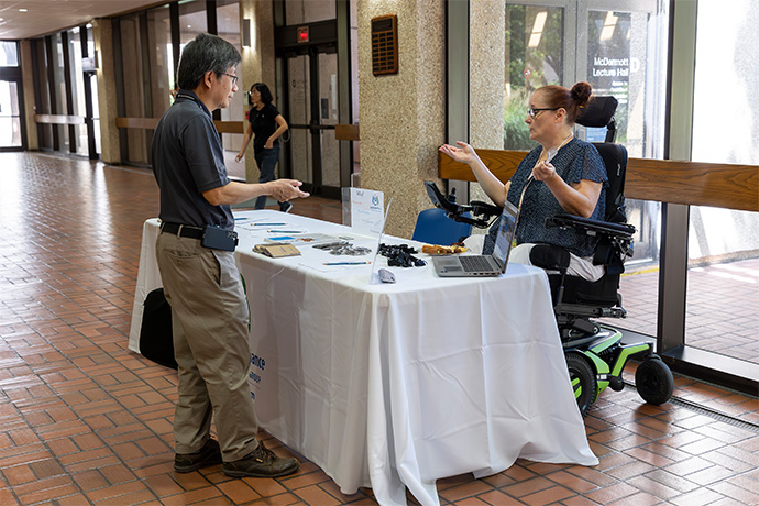 woman in motorized wheelchair wearing glasses and hair in a bun greets male attendee at EquallyAble Alliance BRG event at UTSW