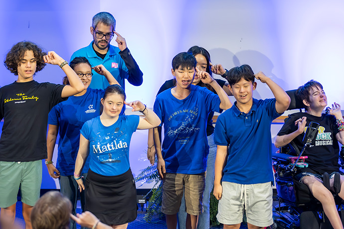 8 diverse performers (age from children to adults, smiling person in wheelchair far right) sign with fingers pointing to their heads on stage at EquallyAble Alliance BRG event