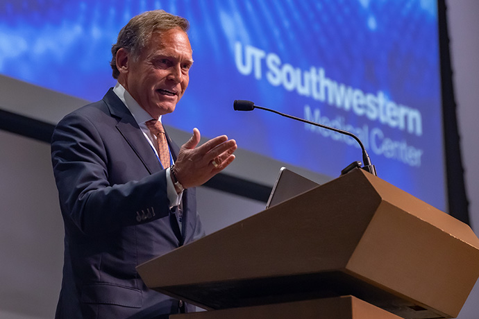 Man with dark, graying hair, wearing a gray suit and glasses, gesturing with his hands as he speaks from a podium. UT Southwestern Medical Center logo is displayed on the screen behind him.