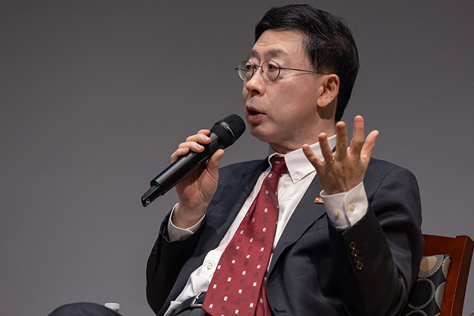 Seated man with dark hair wearing a gray suit, white shirt, red tie, and wire-rimmed glasses speaks into a microphone and gestures with his hands.