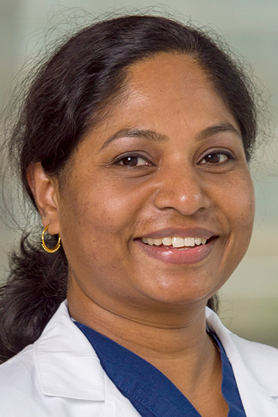 Sydna Sebastian headshot - smiling woman with dark skin, long brown hair in a ponytail, wearing white coat and blue top