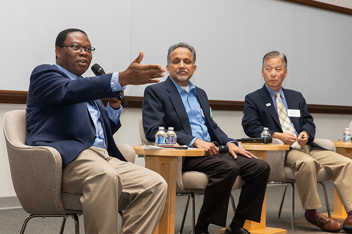 3 men seated on stage as Dr. Achilefu on far left gestures and speaks into microphone, 2 other speakers listen