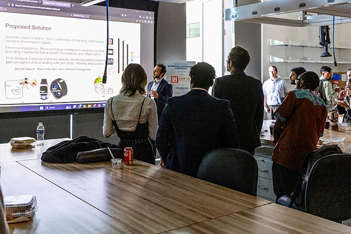 dark meeting room with long table in foreground, 5 people facing forward watch young bearded man in black suit present slide at front of room; slide title: Proposed Solution