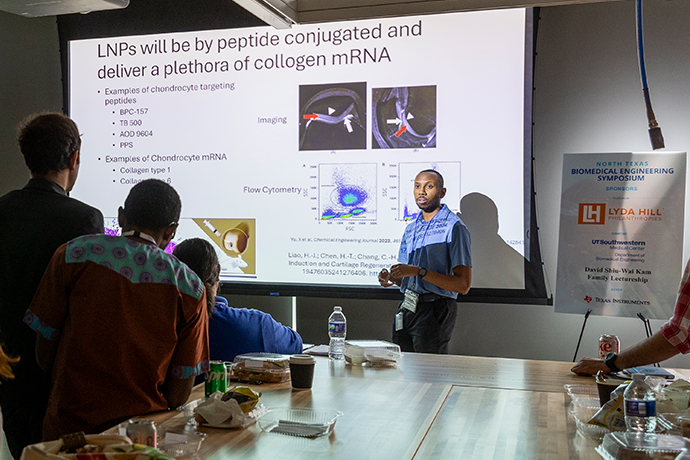 long meeting table in foreground of darkened room; young black man in blue polo shirt stands in light in front of presentation screen showing text and slides: LNPs will be by peptine conjugated and deliver a plethora of collogen mRNA