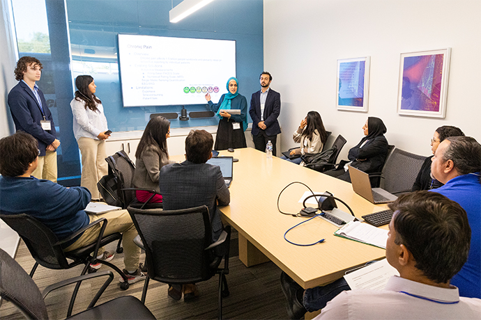 group of 8 diverse people sit around a long table listening to 4 medical students presenting at the front of the room; standing from left: young white man with long dark hair wearing blue blazer, woman with long dark hair wearing grey sweater, woman with blue headscarf and blue dress, dark bearded man wearing black blazer. 