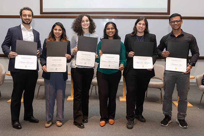 6 diverse people smile and hold award certificates; from left: dark bearded man in black suit with white shirt, short dark-skinned woman with long dark hair, taller woman with black curly hair in gray sweater, dark-skinned woman with shoter dark hair, woman with long black hair in black suit, and dark-skinned man with short hair and glasses wearing black shirt