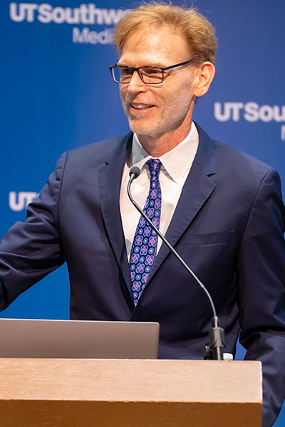 Man with fair hair, wearing glasses and a dark suit, lecturing from a podium.