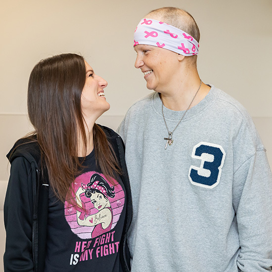 short woman on left with dark hair and black/pink tshirt stands next to cancer patient wife in gray sweatshirt with shaved head and white/pink headwrap