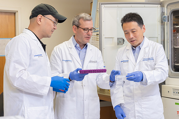 three men in white lab coats and purple gloves look at sample vials in the lab