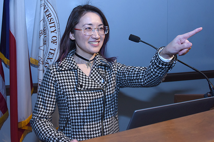 Asian woman with long dark hair wearing black and white plaid jacket speaks from podium, smiling and pointing to audience