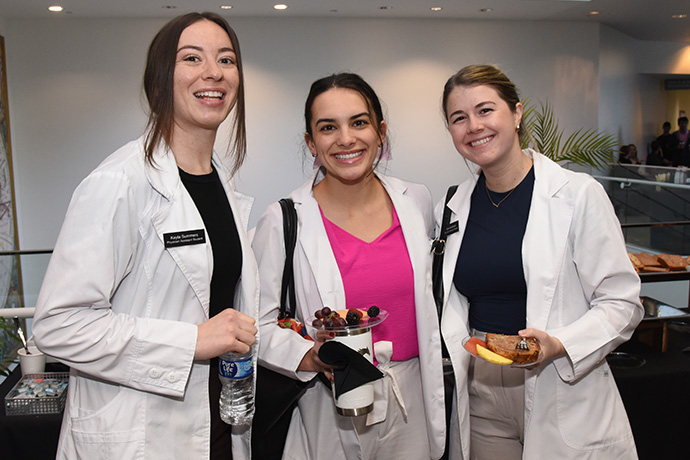 Three smiling woman wearing lab coats posing in front of a buffet table.