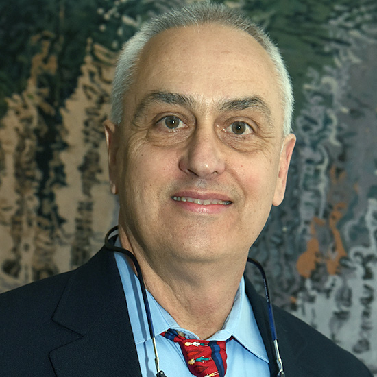 Smiling man with gray receding hair, wearing a dark suit, blue shirt, red patterned tie and a pair of black-framed glasses hanging around his neck.