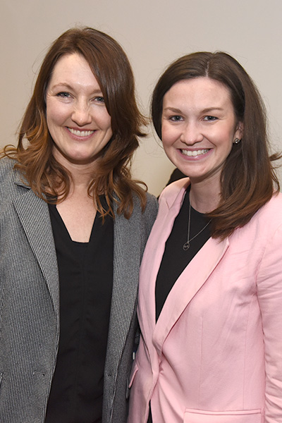 Two smiling women with long brown hair pose for the camera wearing suit jackets over black blouses. One jacket is gray and the other is pink.